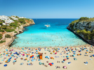 Aerial view of colorful umbrellas on crowded sandy beach, boats, hotel, rocks, swimmers in blue sea, green trees on sunny summer day. Mallorca, Balearic Islands, Spain. Tropical top drone view. Travel