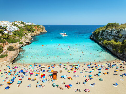 Aerial view of colorful umbrellas on crowded sandy beach, boats, hotel, rocks, swimmers in blue sea, green trees on sunny summer day. Mallorca, Balearic Islands, Spain. Tropical top drone view. Travel