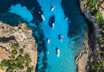 Aerial top view from drone of yachts and boats floating in turquoise sea between rocky cliffs, swimmers and paddleboards enjoying sunny summer day in Mallorca, Balearic Islands, Spain. Seascape