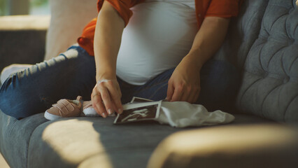 Fototapeta premium Pregnant woman holding ultrasound scan with baby clothes and shoes