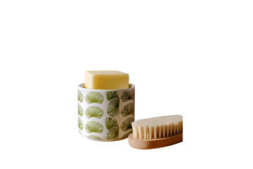 A white, round container with green patterns on the side, containing a yellow soap bar. Next to it is a pink, knitted cloth and a wooden brush isolated on a transparent background in PNG format.