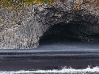 Hexagonal basalt columns lead into a dark cave on a black sand beach. Ocean waves meet the shore, with greenery atop the rock formation.