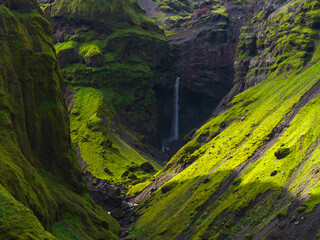 A narrow waterfall cascades into a shadowy Mulagljufur gorge surrounded by steep moss covered cliffs, with sunlight highlighting the vibrant green terrain. © Aerial Film Studio