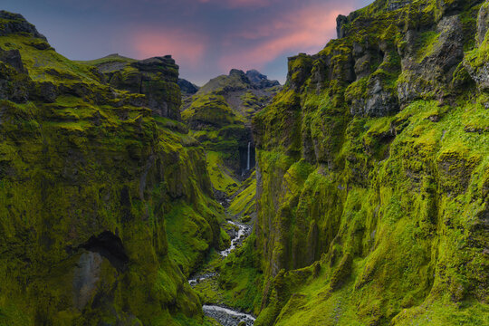 Mulagljufur Canyon in Iceland features moss covered cliffs, a winding river, and a distant waterfall under a pink and purple sunset sky. - Powered by Adobe
