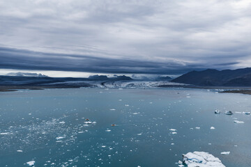 Aerial view of Jokulsarlon Glacier Lagoon in Iceland, featuring blue waters with icebergs, a massive glacier, dark mountains, and a small boat.