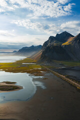 Aerial view of Stokknes Peninsula in Iceland, showing jagged mountains, black sand, a reflective water body, a road, and patches of green vegetation.
