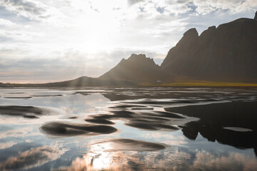 The Stokksnes Peninsula features the Vestrahorn mountains, a black sand beach, and shallow water pools reflecting the sky under soft sunlight.
