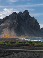 Fototapeta premium The Stokksnes Peninsula features Vestrahorn mountain's jagged peaks, a black sand beach with green grass, and ocean waves under a partly cloudy sky.