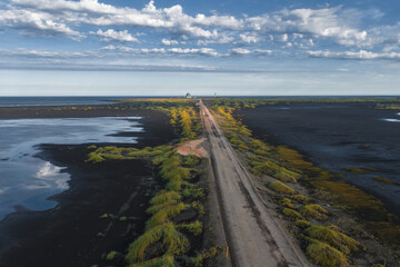 A straight dirt road crosses a black sand terrain with green vegetation. A lighthouse or tower and a small structure are visible under a partly cloudy sky.