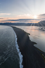 Fototapeta premium Aerial view of a black sand beach in Iceland with ocean waves, calm reflective waters, and a distant mountain range under soft sunlight.