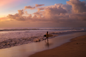 Naklejka premium A young surfer walks along Seminyak Beach, Bali, after training, holding a surfboard. The slightly blurry photo adds a dramatic, dreamy effect to the sunset-lit waves and sandy shoreline.