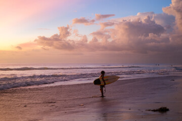 A young surfer walks along Seminyak Beach, Bali, after training, holding a surfboard. The slightly blurry photo adds a dramatic, dreamy effect to the sunset-lit waves and sandy shoreline.