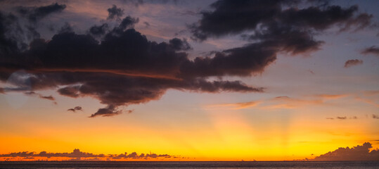 Fiery sunset over the ocean paints the sky with glowing orange, gold, and purple tones, while dark clouds drift across the horizon in a dramatic evening scene.