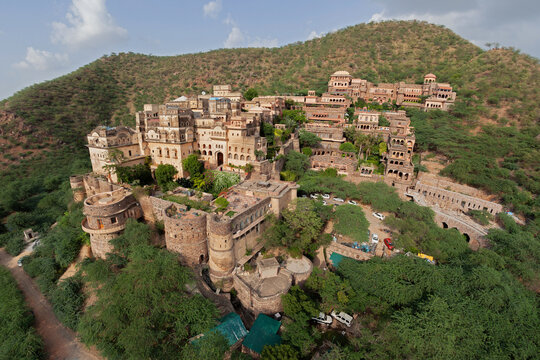 Aerial view of the ancient fort complex nestled on the hillside, its stone walls a tapestry of ochre and umber against the vibrant green foliage, Rajasthan, India.