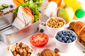 School healthy food for children. Preparing homemade balanced healthy lunch box for schooler, with sandwich, fruits, berries, juice, nuts. Ingredients and foods with lunch box on white table
