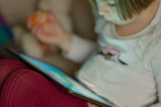 A little girl wearing a colorful mask on her face is happily using a tablet computer, deeply engaged in her learning activity - Powered by Adobe
