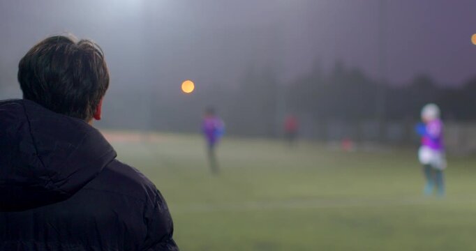 A man watches a soccer game on a foggy night, with the field illuminated and shadows across the grass, creating a mysterious atmosphere - Powered by Adobe