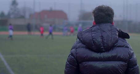 A man in a black jacket watches an exciting soccer game from the sidelines, observing the spirited play of children in a friendly match