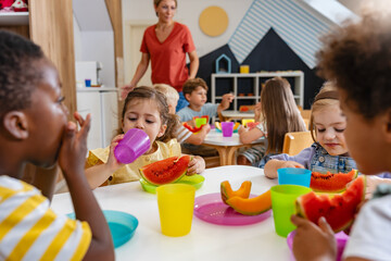 Preschool children eating fresh watermelon slices at table, enjoying healthy snack in bright classroom