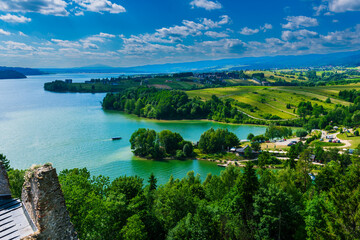Elevated view of Czorsztyn Lake with a green peninsula, trees, and a tourist boat. Rolling hills and farmland extend into the distance.