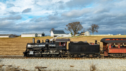 A vintage steam locomotive moves along railroad tracks across a scenic rural area. The backdrop features expansive fields and distant barns under cloudy skies, evoking a sense of nostalgia.