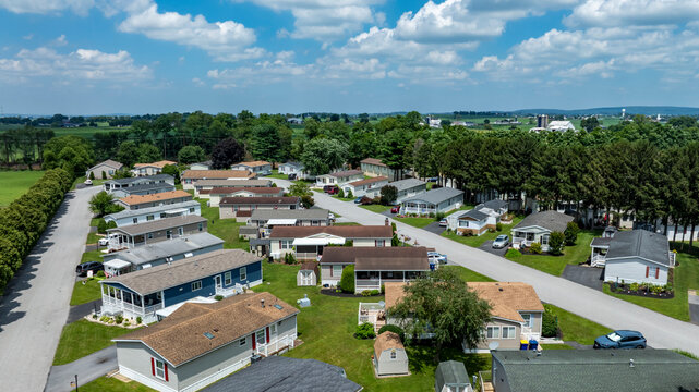 A peaceful community features rows of mobile homes surrounded by lush greenery under a bright blue sky. The setting is calm, showcasing well-maintained yards and roads.