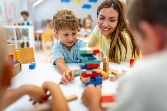Smiling preschool boy playing with colorful wooden blocks, guided by a cheerful teacher, in a bright and lively classroom with other children in the background