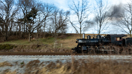 A classic steam locomotive speeds along a gravel track in a scenic rural area. Trees line the route as smoke billows from the engine, creating a nostalgic atmosphere.