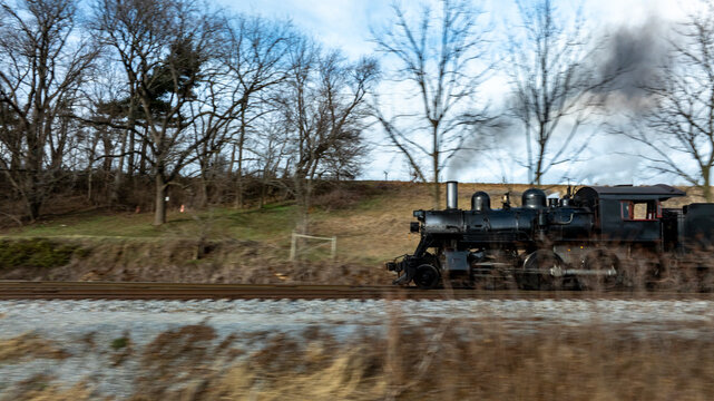 A classic steam locomotive speeds along a gravel track in a scenic rural area. Trees line the route as smoke billows from the engine, creating a nostalgic atmosphere. - Powered by Adobe
