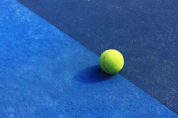 A single tennis ball sits on a blue court with two different shades, ready for a game, match, or tournament, under bright lighting