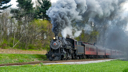 A vintage steam locomotive emits dense black smoke while moving along a scenic railway surrounded by lush trees.