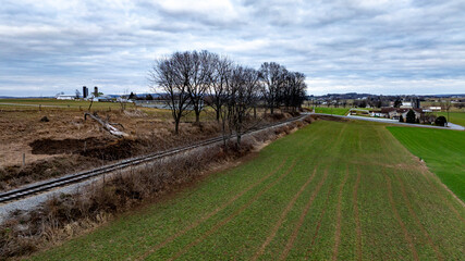 Wide green fields stretch beside weathered railway tracks in a rural area. Leafless trees stand tall against the cloudy sky, creating a tranquil atmosphere in this scenic countryside setting.