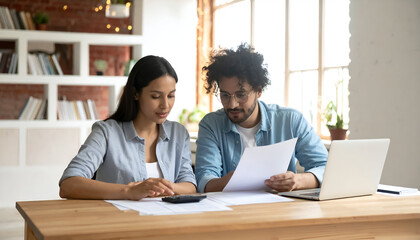 Young couple calculating finances at home on wooden desk with laptop and papers, feeling stressed about bills and budget, in natural light and modern interior.