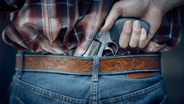 A cowboy is pulling a gun from his waistband to protect himself from enemies. A country cowboy man embodies the cowboy.