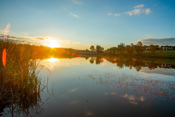 A tranquil evening view of a calm lake, with a bright sunburst and lens flares over the water, and a green shoreline with a distant treeline