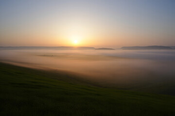 Golden sun rising above a thick blanket of morning mist covering a picturesque green landscape.