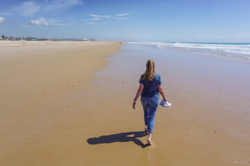 A barefoot woman walks along the shore of a long golden sand beach with the blue sea in the background
