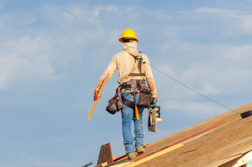 Construction worker in safety harness and hard hat walks on roof frame carrying a measuring square and nail gun, representing residential roofing and carpentry work
