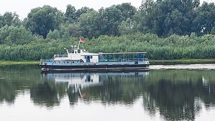 Barge cruising along a calm waterway surrounded by lush greenery during a cloudy day in a rural landscape
