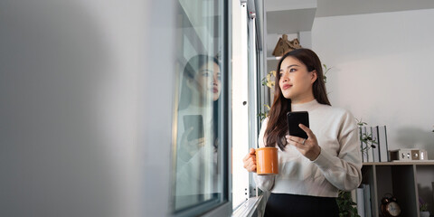 Thoughtful woman holding coffee and smartphone, gazing out window