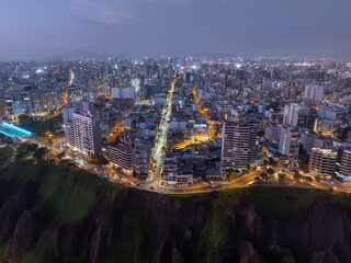 Aerial night view of Miraflores district in Lima, Peru, showing illuminated streets, modern buildings, and the coastline cliffs along the Pacific Ocean.
