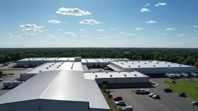 Aerial drone view of a large modern industrial warehouse and distribution center complex with loading docks, set against a blue sky and surrounded by a vast green forest.