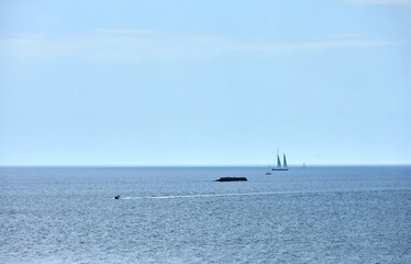 A motorboat and sailboats navigate the open sea near a small rock formation under a bright blue sky, evoking a peaceful and minimalist atmosphere. boating, minimalism