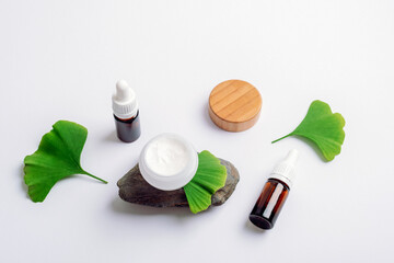Glass jars with dropper bottles, green ginkgo leaves and face cream on stone on white background. Natural skincare and wellness concept
