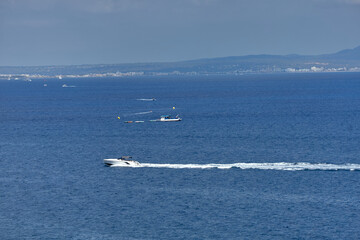 Obraz premium A white speedboat and another vessel tow a water tube across the open sea near a distant coastline under a cloudy sky. water recreation