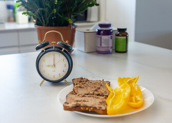 Close up of pâté sandwiches with yellow pepper on white plate, alarm clock and vitamins, symbolizing breakfast, healthy lifestyle and daily planning