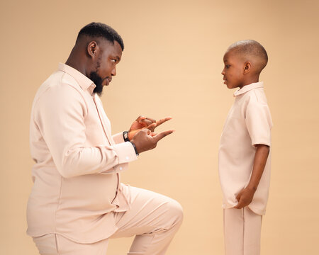 Nigerian father and son in matching beige exchange a playful spider hand sign, their smiles capturing joyful family bonding, love, tradition, and a lighthearted moment together. - Powered by Adobe
