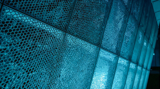 A close-up, abstract view of a curved wall with a repeating hexagonal pattern and textured glass panels, illuminated with a cool blue light.