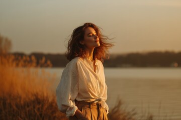 Relaxed woman enjoys sunset with closed eyes near lake and grass field. Concept for mindfulness retreat, tranquil vacation and outdoor lifestyle promotion