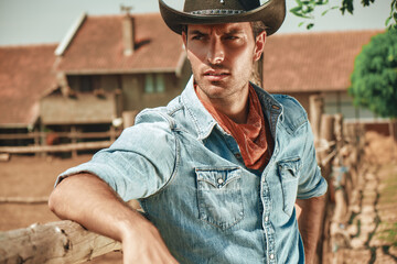 A young cowboy leans against a wooden fence at a ranch, wearing a denim shirt and cowboy hat. The golden hour light enhances the warm tones of the rural landscape behind him